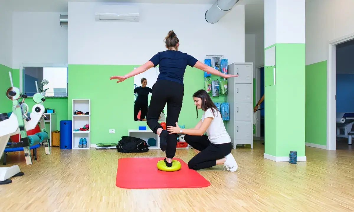 Kobieta ćwiczy propriocepcję na poduszce balansującej, druga osoba asekuruje.