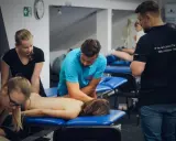 Uczestnicy szkolenia wykonują masaż tkanek miękkich na pacjencie leżącym na kozetce. Terapeuta w niebieskiej koszulce demonstruje technikę.