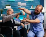 Starszy mężczyzna w wózku wykonuje ćwiczenia z hantlem pod okiem fizjoterapeuty. To kluczowy element rehabilitacji ręki po udarze.