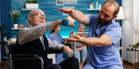 Starszy mężczyzna w wózku wykonuje ćwiczenia z hantlem pod okiem fizjoterapeuty. To kluczowy element rehabilitacji ręki po udarze.