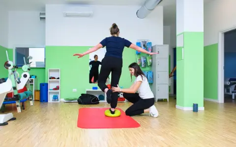 Kobieta ćwiczy propriocepcję na poduszce balansującej, druga osoba asekuruje.