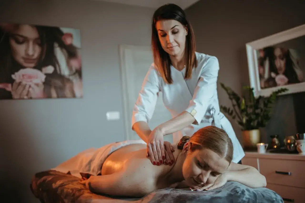 Kobieta otrzymuje masaż relaksacyjny plec&oacute;w. Terapeuta w białej bluzie delikatnie ugniata jej ramiona.