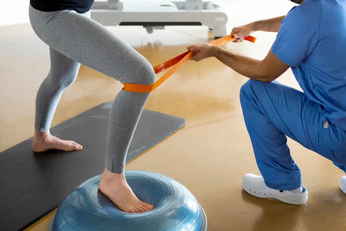 Ćwiczenia z taśmą oporową na piłce bosu pomagają jak wzmocnić kolana. Fizjoterapeuta asystuje pacjentowi.